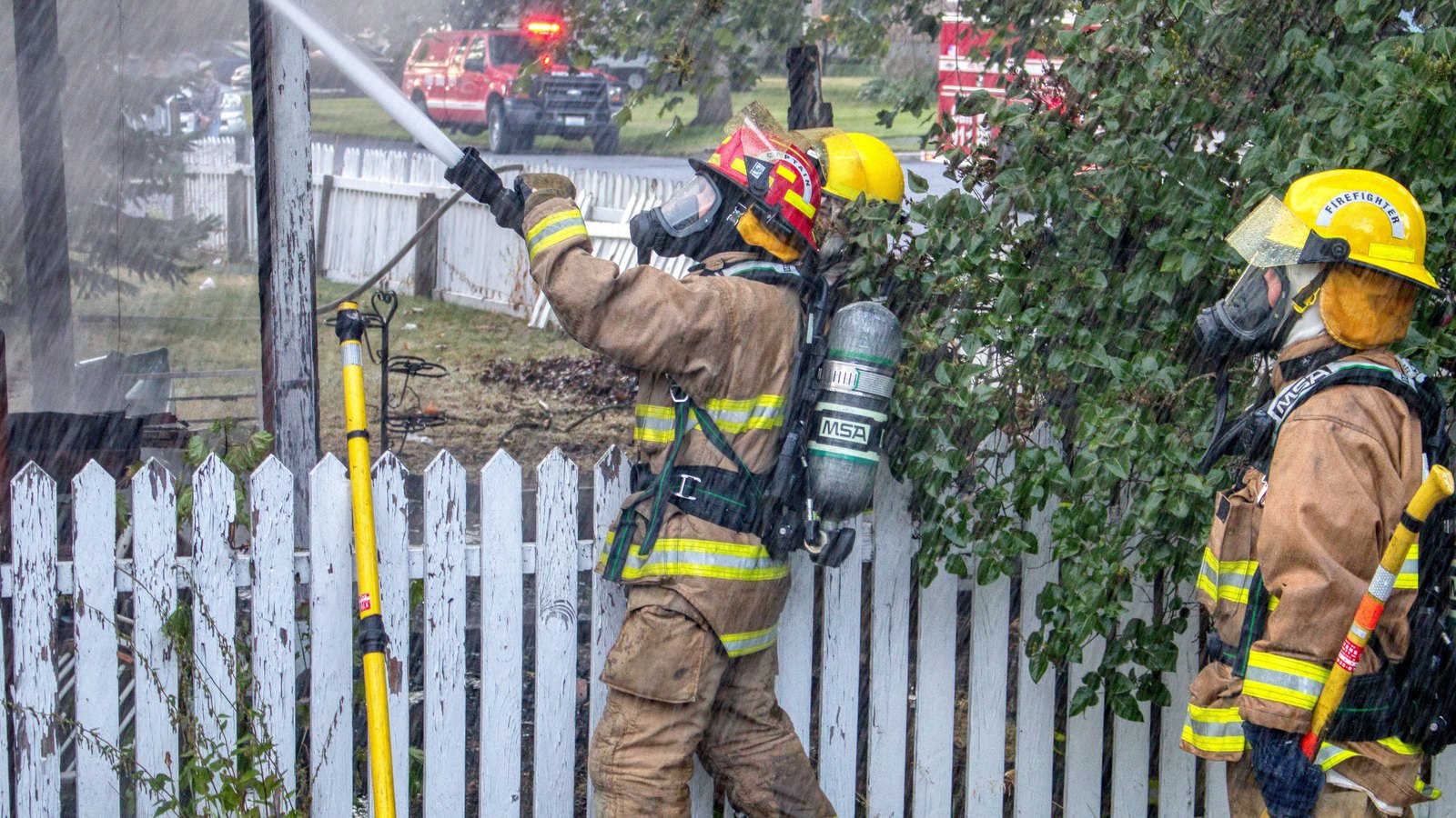 Two firefighters in tan turnout gear and yellow helmets aim a hose stream over a white picket fence toward a house, a red fire engine blurred in the street behind them. No one is identifiable and no signage, plate or landmark places the scene. It illustrates a residential firefighting response and is not a photograph of this fire or of Scenic Street.