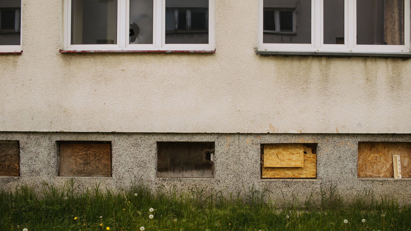 A weathered rendered wall under flat grey light, its upper windows empty with one pane broken, and a row of low openings along the base sealed with sheets of raw chipboard, one of them left open and darkened with soot. Long uncut grass and weeds run across the whole foot of the wall. No person is in frame and no house number, sign, lettering or agency mark is legible anywhere. Stock photo, not a photograph of this event.