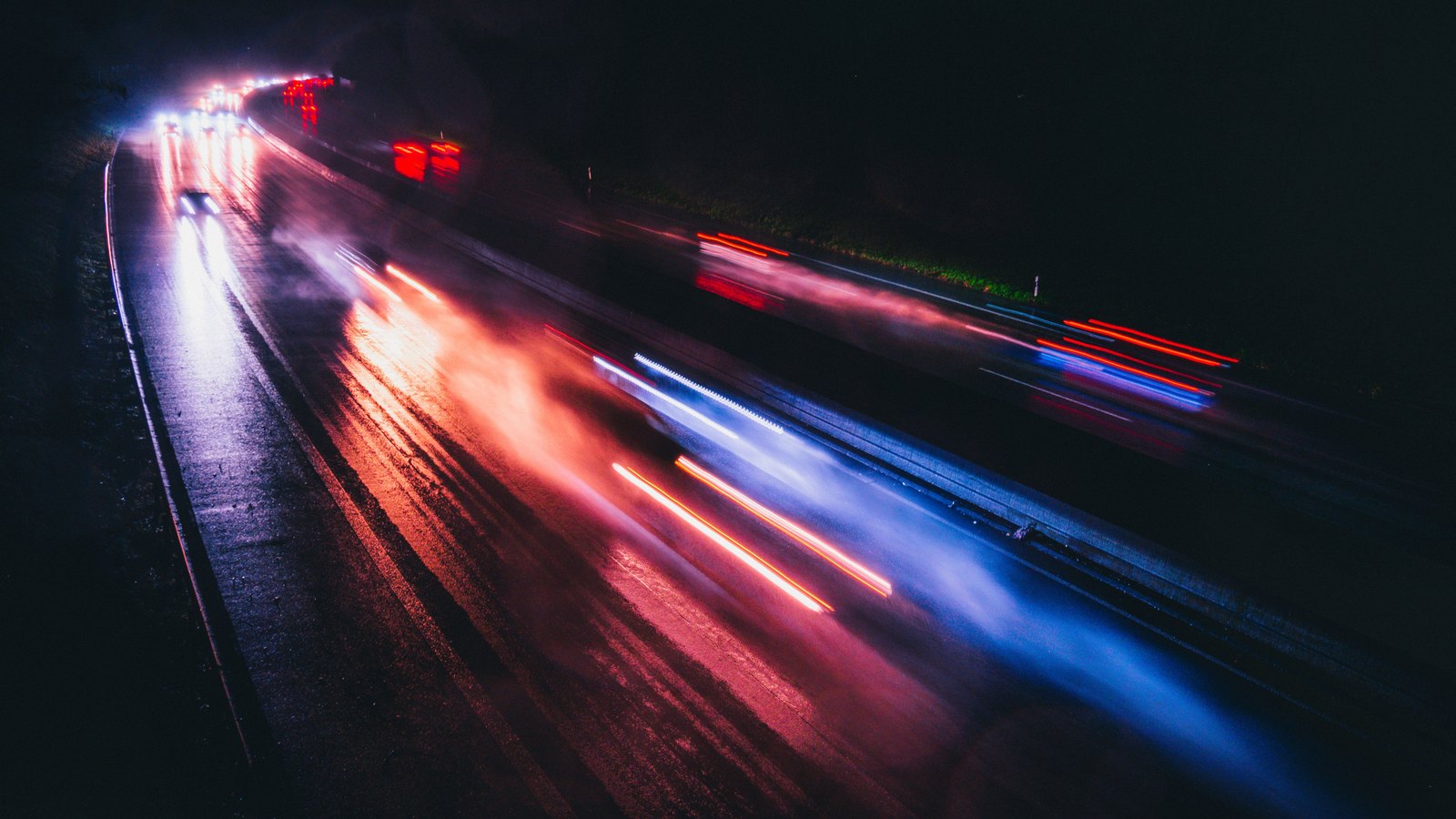 Red and blue light streaks smear across a wet, dark road at night in a long exposure, seen from above the carriageway. No person, vehicle, signage, plate or landmark can be made out. It illustrates a night road incident with emergency lights and is not a photograph of this crash or of Sunshine Boulevard.