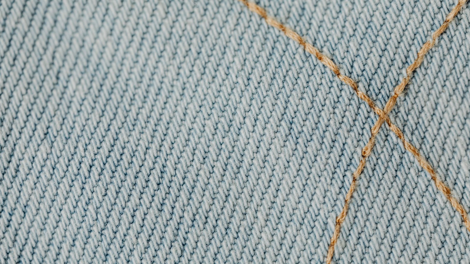 A close photograph of pale blue denim, the diagonal twill of the weave running corner to corner and two lines of tan topstitching crossing near the right of the frame. No text, brand mark, label, person or place is in view.