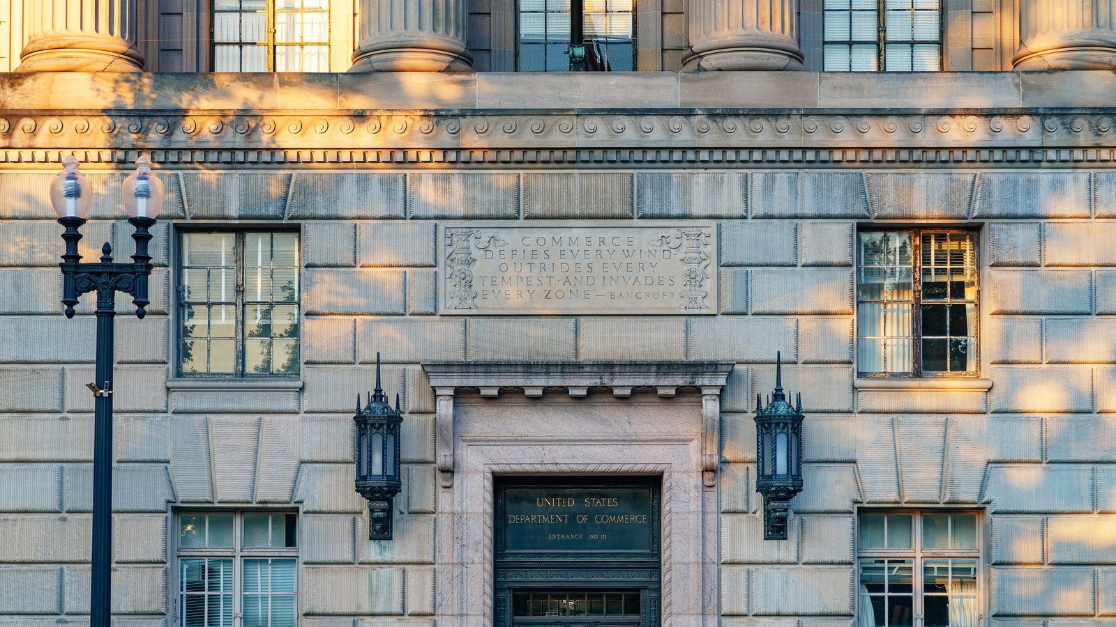 The limestone front of the Herbert C. Hoover Building in Washington at low sun, a bronze door marked United States Department of Commerce, Entrance No 10, set beneath a carved panel reading Commerce defies every wind, outrides every tempest and invades every zone, with two iron lanterns either side and tree shadows across the masonry.