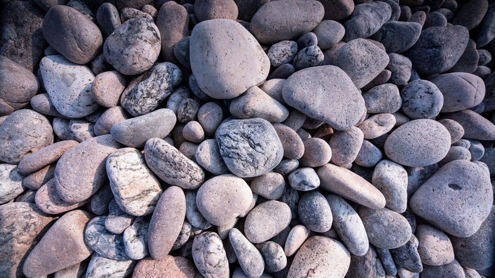 Smooth rounded stones in grey, pink and cream are packed tightly against each other and fill the whole frame, several flecked or veined with darker mineral, each catching a soft highlight along its upper edge. No text, brand mark, person, water or horizon is in view.