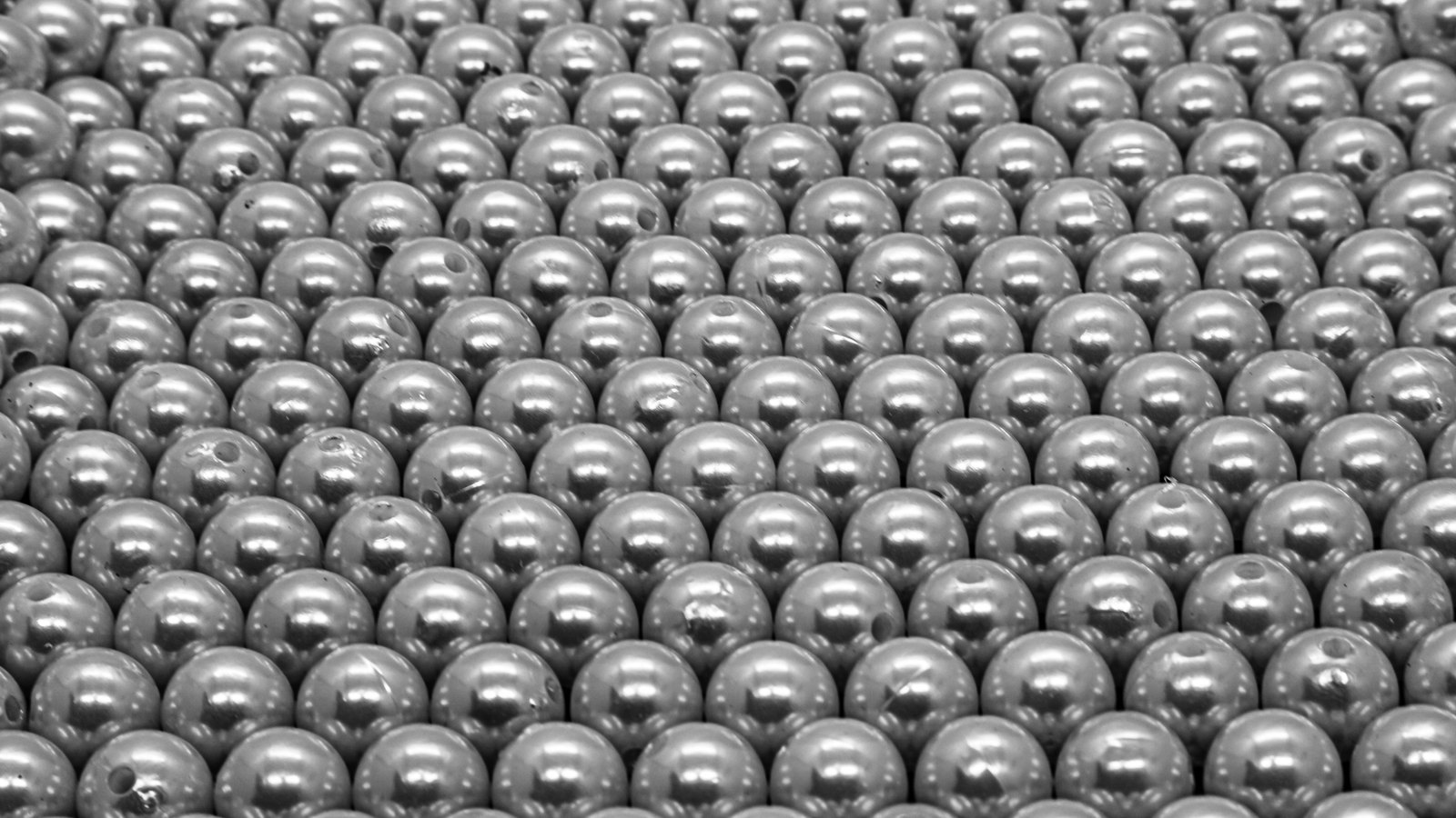 A dense field of pale grey spherical beads packed edge to edge in tight rows, filling the entire frame and receding slightly towards the top. Each bead carries a hard white highlight, and several show a small dark drilled hole. No text, brand mark, person, place, sky or background surface is in view.