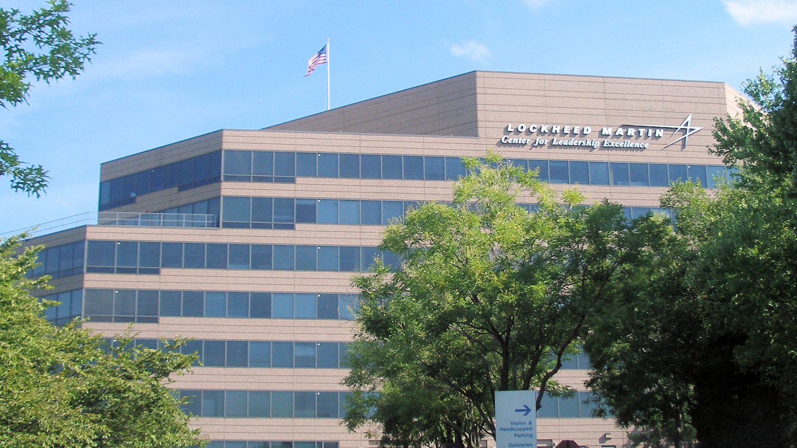 A low angle view through summer trees of a stepped office building faced in pale pink stone with bands of dark tinted glazing, carrying the name Lockheed Martin and the line Center for Leadership Excellence in metal letters near the roofline, with a United States flag flying from a mast above the roof against a blue sky.
