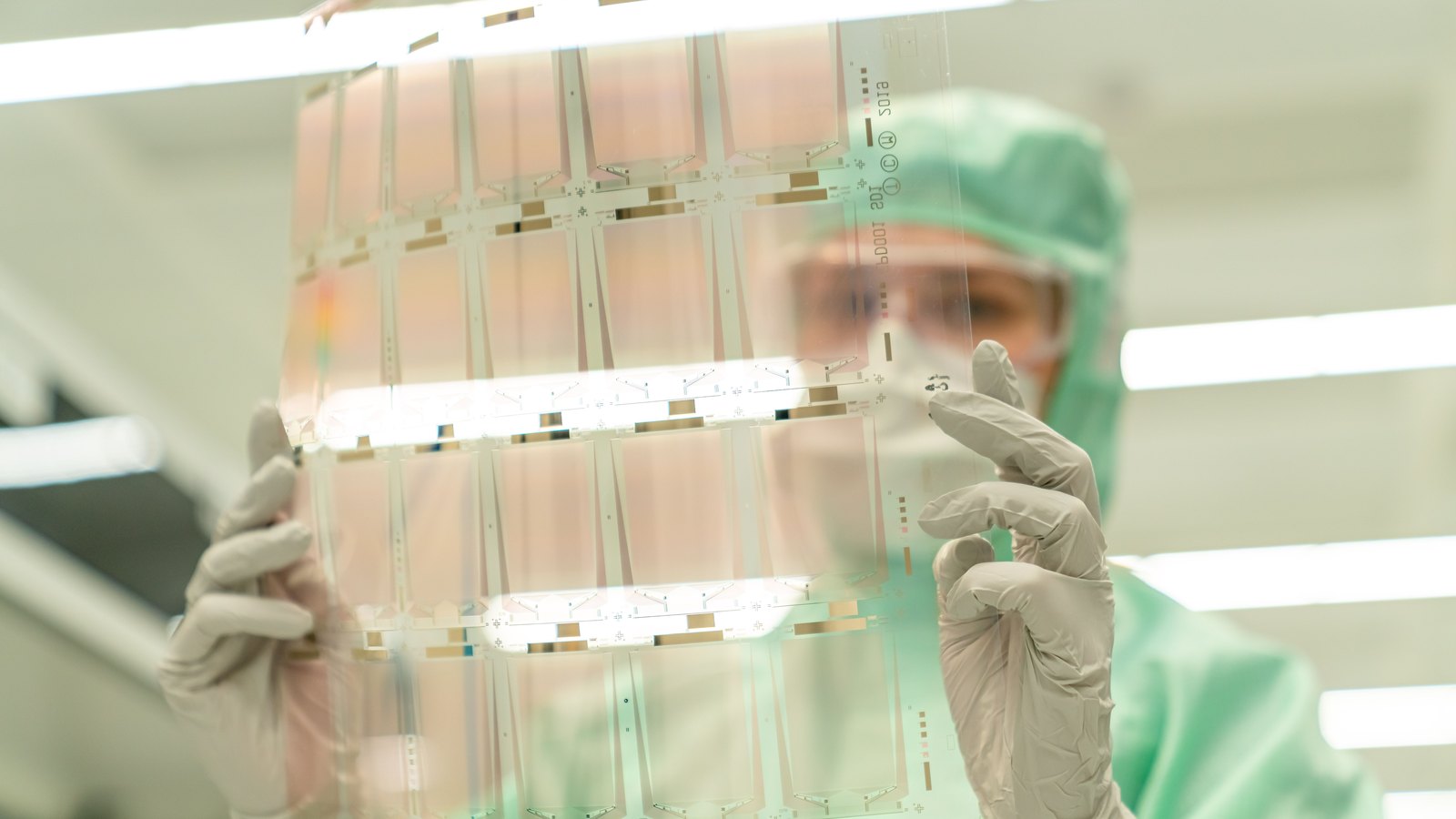 A cleanroom technician examines a transparent electronics sheet under bright laboratory lights.