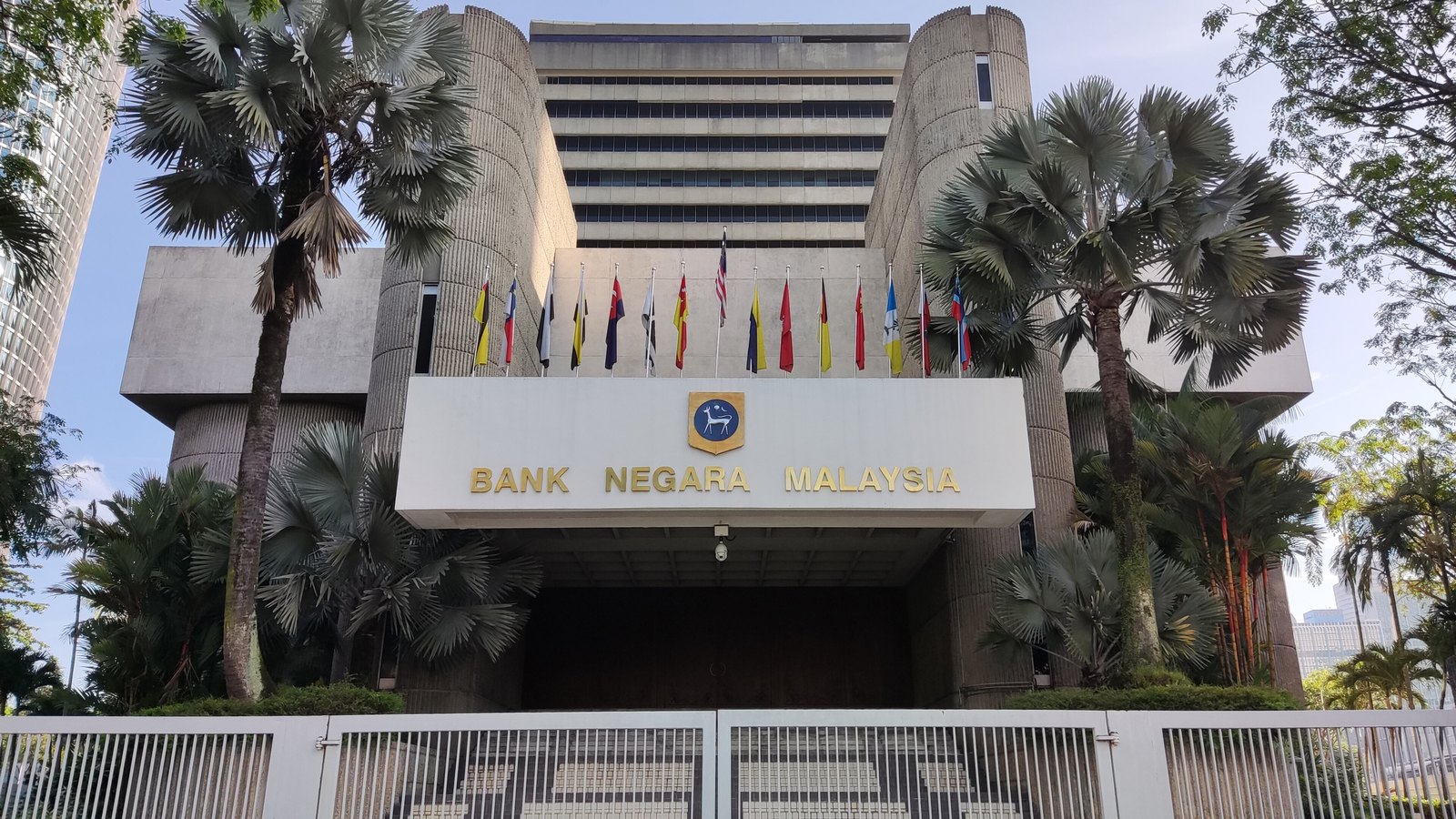 The Bank Negara Malaysia headquarters in Kuala Lumpur, framed by palm trees and national and state flags.