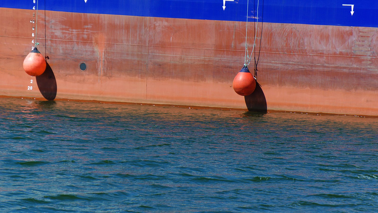 The steel side of a large ship at the waterline filling the frame: red oxide hull plating below a blue topside band, white draft numerals painted in a vertical column towards the left, load line arrow marks at the top right, and two orange pneumatic fenders hung on lines against the plating. Calm green water fills the lower half of the frame. No vessel name is in view.