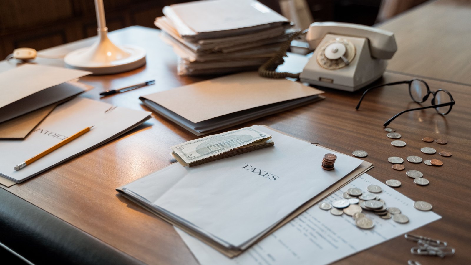 A wooden desk photographed at an angle, with a paper folder marked TAXES lying open at the centre, a small folded stack of United States banknotes and a short pile of copper coins resting on it. Loose silver and copper coins are scattered across a printed sheet to the right, beside a pair of round spectacles. Behind them sit a stack of buff document folders, a cream rotary dial telephone, a pencil, a pen and the base of a desk lamp. No person, no signage, no place name and no ballot is in view.