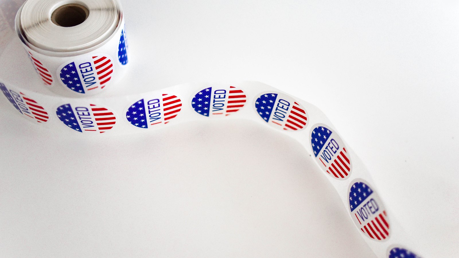A long strip of round I VOTED stickers, red and blue with a stars and stripes design, unspooling from a roll in the top left corner and curving away across a plain white surface. No person, no place, no building and no agency name appears in the frame. Stock photo, not a photograph of this event.