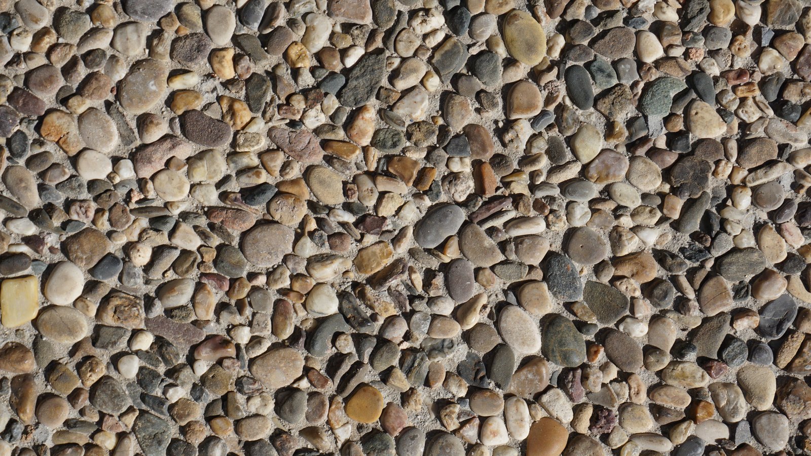 An exposed aggregate concrete surface photographed straight on and filling the whole frame, packed with hundreds of small rounded pebbles in grey, tan, cream and rust tones set into pale cement. No horizon, no sky, no person, no text, no logo and no identifiable place appears in the image.