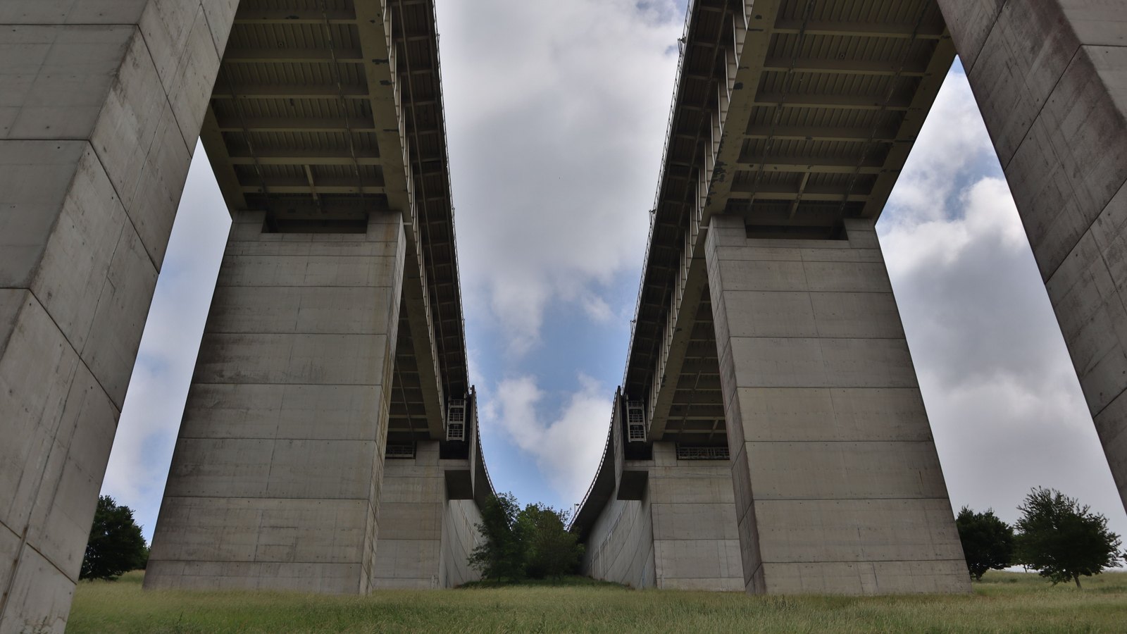 The underside of a tall concrete road viaduct seen from the grass below, its two decks curving away into the distance between massive square piers.