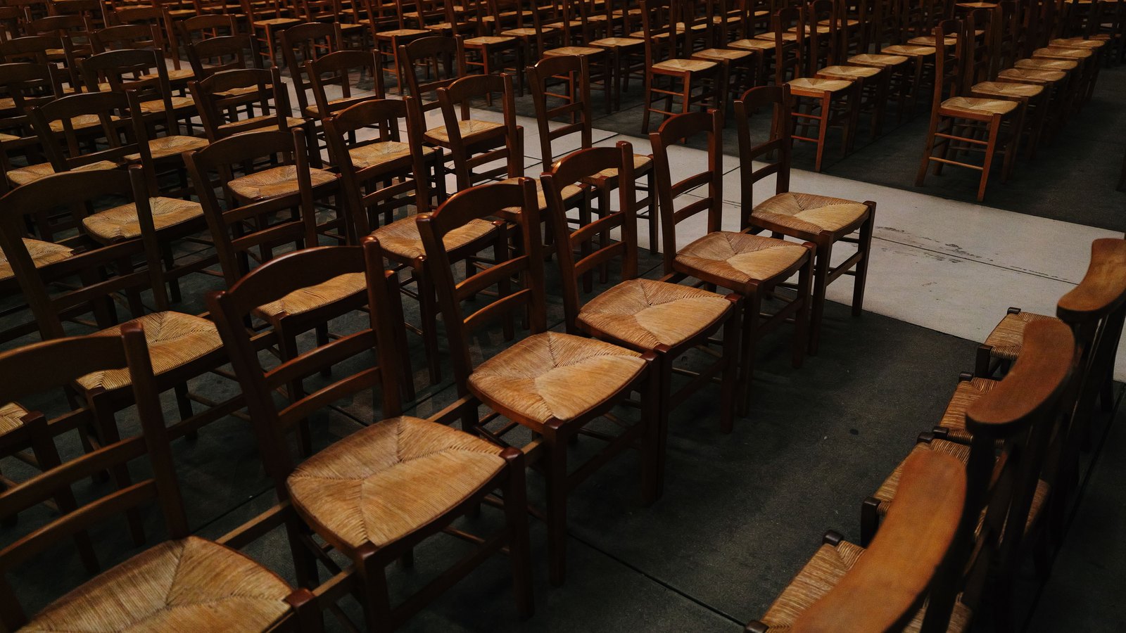 Rows of empty dark wooden chairs with woven rush seats, set out in long lines across the bare concrete floor of a dim hall and photographed from above and to one side. A pale strip of flooring runs between two blocks of seating like an aisle. Every chair is unoccupied. No person, no signage, no place name and no ballot is in view.