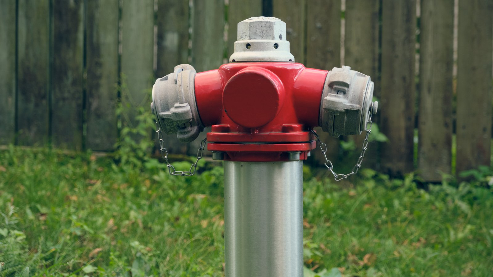 A red fire hydrant standing in grass in front of a weathered wooden fence, photographed head on in flat daylight, with no people, vehicles, buildings, signs or text in view.