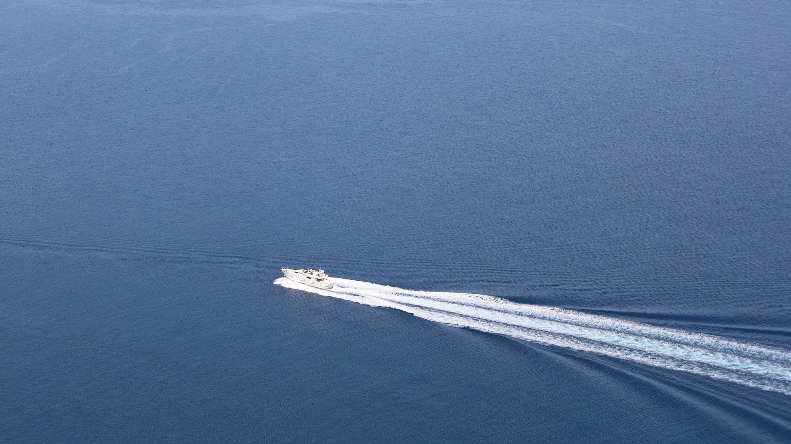 A high aerial view of a single white motor vessel running alone across flat, deep blue open water, trailing a long white wake that reaches to the edge of the frame. There is no coastline, no horizon and no other vessel in sight. No boat name, registration number, flag, agency livery, person's face or identifiable place appears anywhere in the frame. A generic illustration of a boat alone on open water, not a photograph of this event or of the vessel involved.