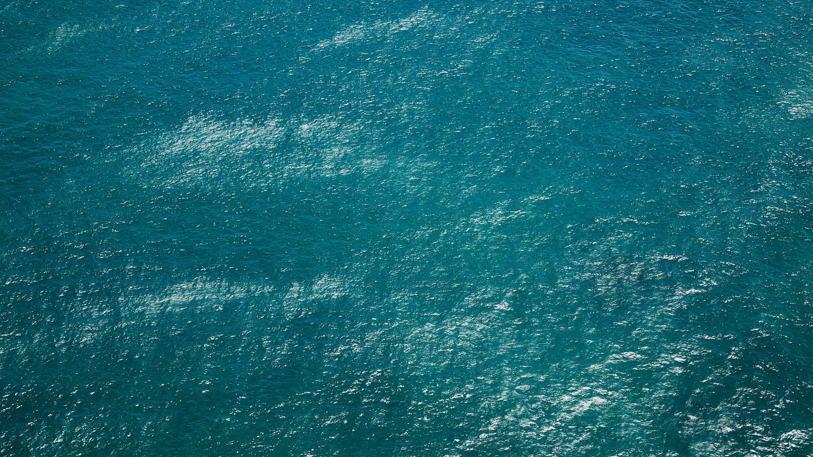 An overhead crop of open turquoise water filling the frame, its surface broken into small ripples with scattered pale glints of reflected sunlight. There is no horizon, shoreline, vessel, person, structure or legible text in view, and no location can be identified from it. An abstract water texture used as a plain illustration, not a photograph of the Inlet Drive property or of this fire.