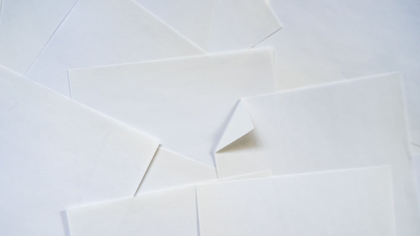 A dozen blank white sheets of paper scattered in overlapping layers across a white surface, filling the frame, with soft grey shadows along their cut edges and one corner turned up near the centre. Every sheet is unprinted. No text, no person, no logo and no identifiable place appears in the image.