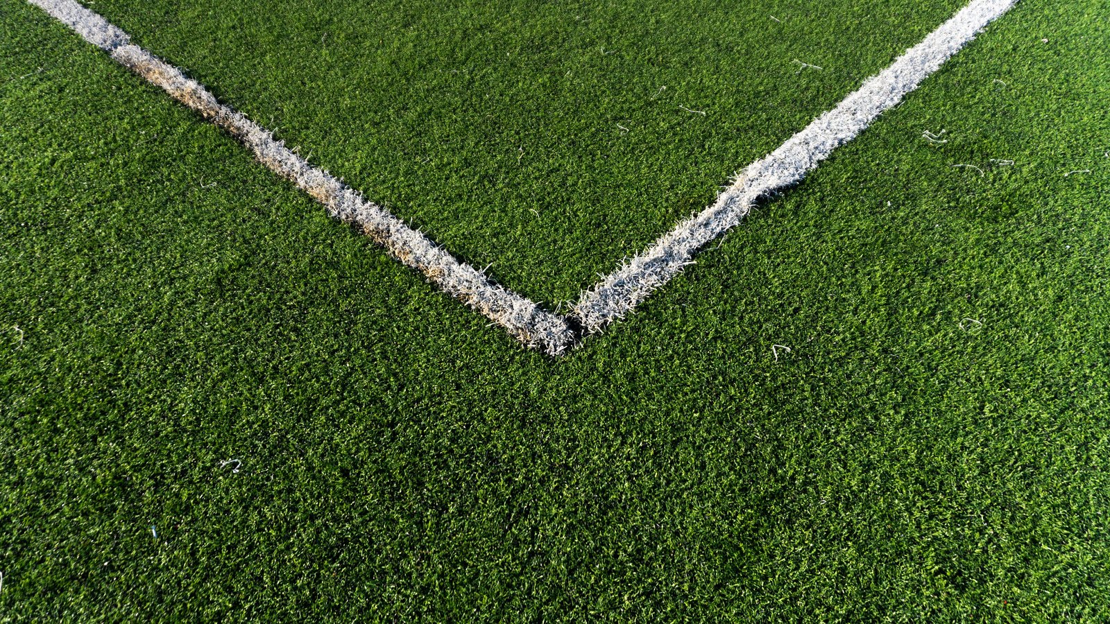 A close overhead photograph of green artificial turf, with two white painted boundary lines meeting at a corner near the centre of the frame and running off to the top left and top right. The paint is worn and flecked and a few loose fibres lie on the surface. No people, buildings, equipment, signage, lettering or brand marks are visible, and no place or school can be identified from it. A generic illustration of a playing surface, not a photograph of Mariner High School.