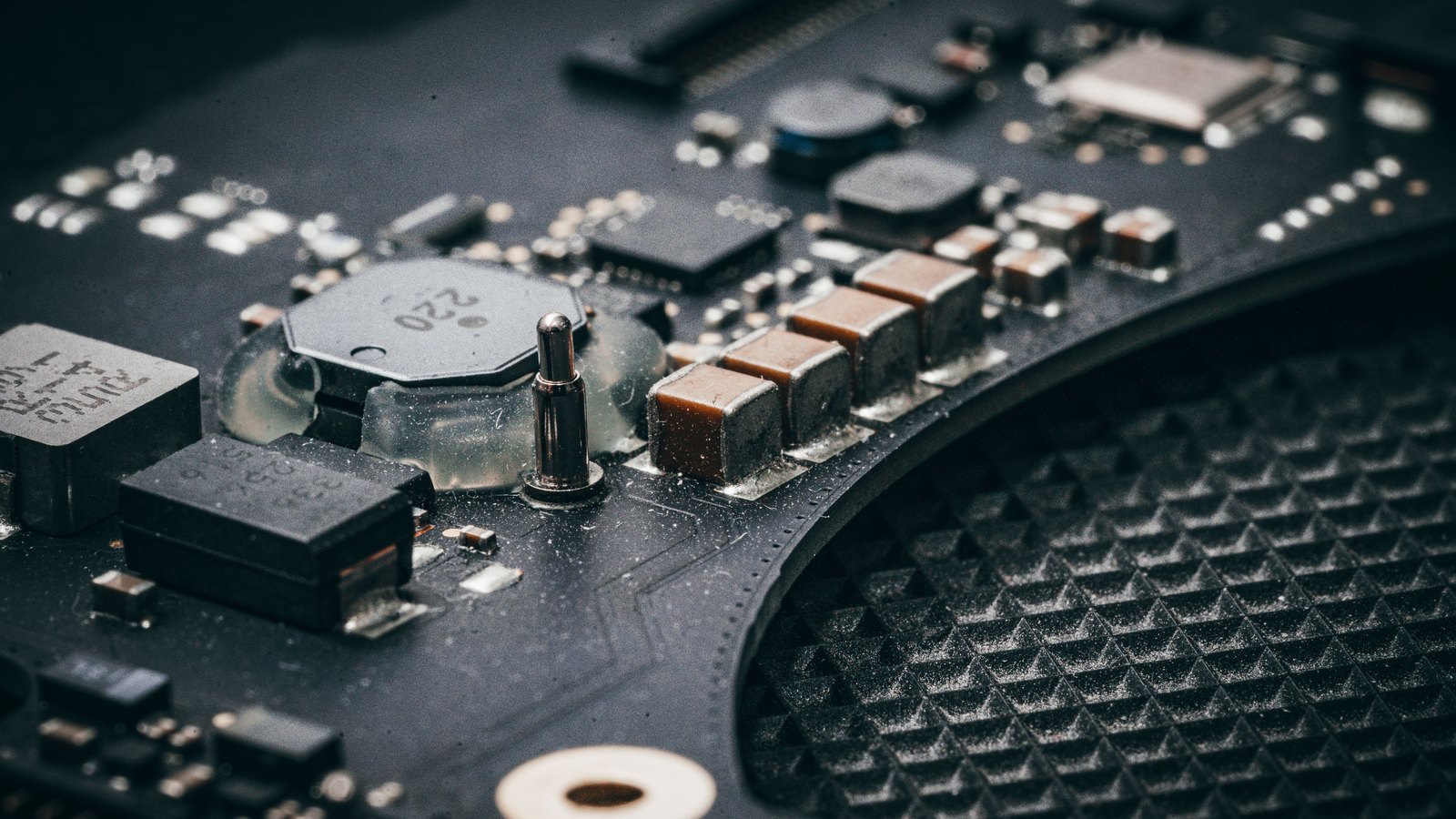 A dusty macro view of a dark circuit board, with a coin cell, a row of copper coloured capacitors, a small metal pin and scattered surface mount components picked out in low side light.