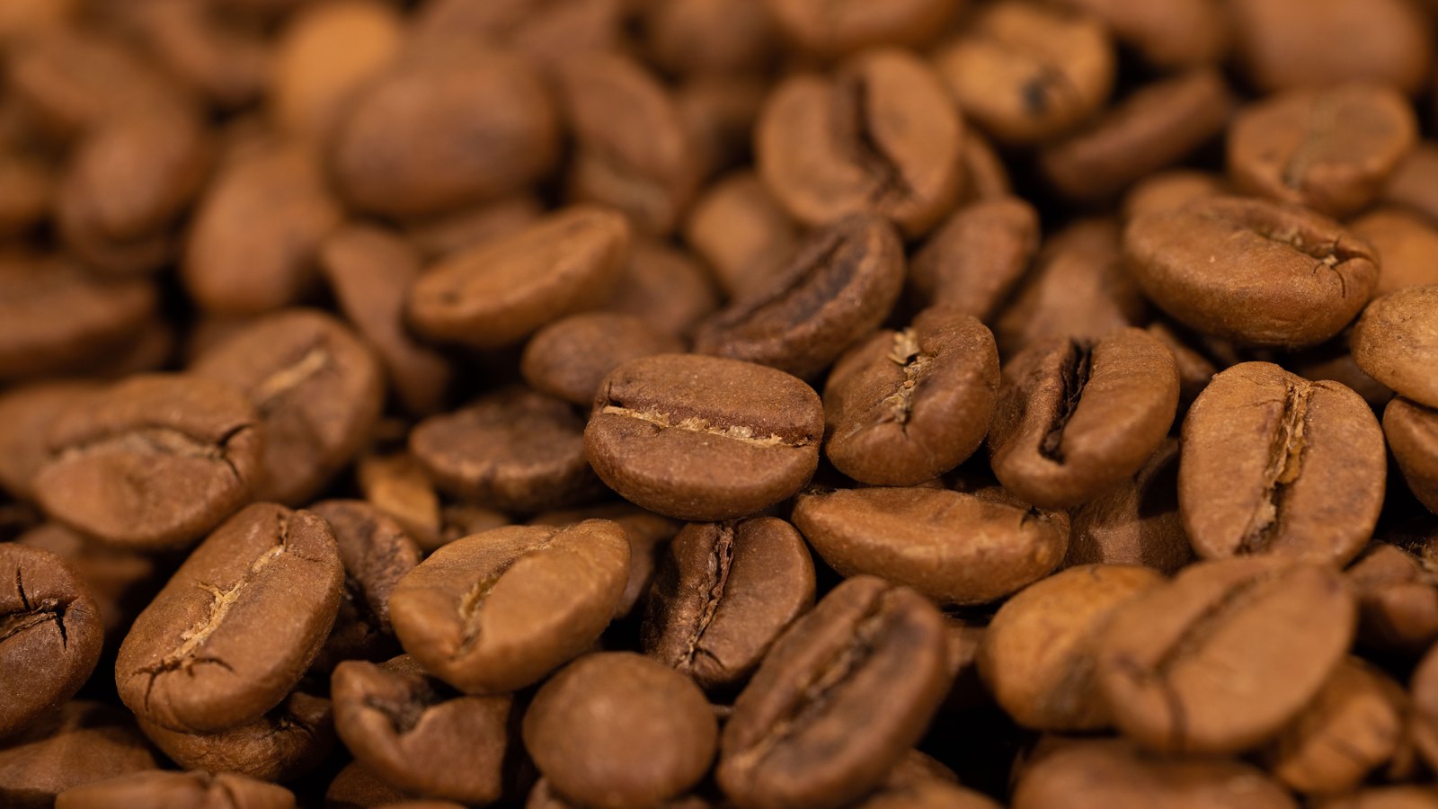 A tight macro of roasted coffee beans heaped so that they fill the whole frame, each bean showing the pale crease down its centre against a warm brown surface, with the beans at the top and bottom edges softening out of focus. No packaging, person, lettering or brand mark is visible.