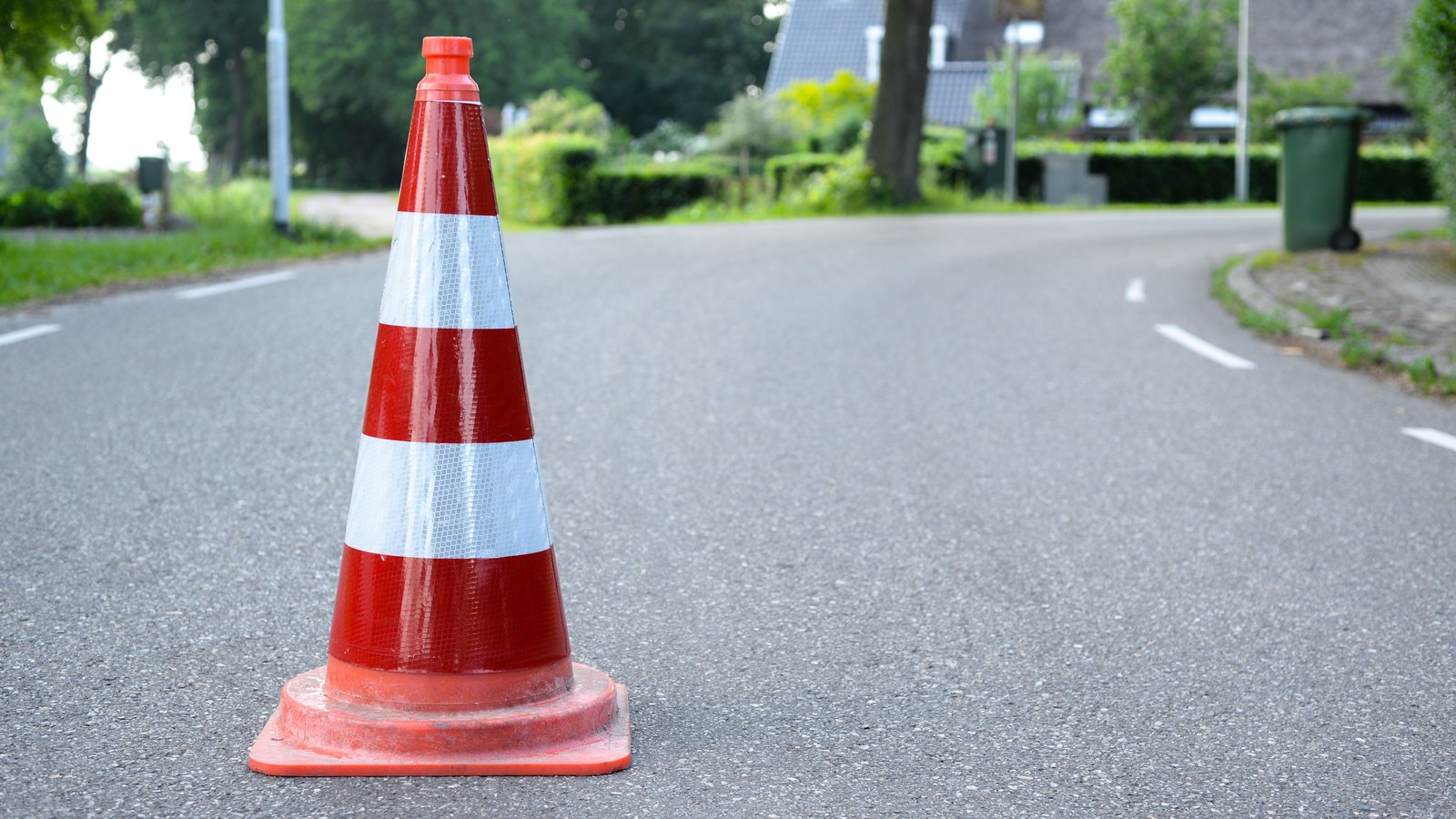 A single red and white traffic cone standing in close foreground on open grey asphalt, with a quiet residential road curving away behind it into soft focus past a clipped hedge, a house roof and a green wheeled bin. There is no text, no signage, no lettering, no brand mark, no person and no vehicle anywhere in the frame, and no landmark that identifies any place. A generic illustration of a road closure, not a photograph of McGregor Boulevard, of Bay Drive, of any downed power line or of anything in Lee County. Two weaknesses are stated rather than glossed: a traffic cone is a generic closure symbol and this closure is by power lines down across the road with traffic being turned away, not a coned off lane, and the leafy background is not Southwest Florida.