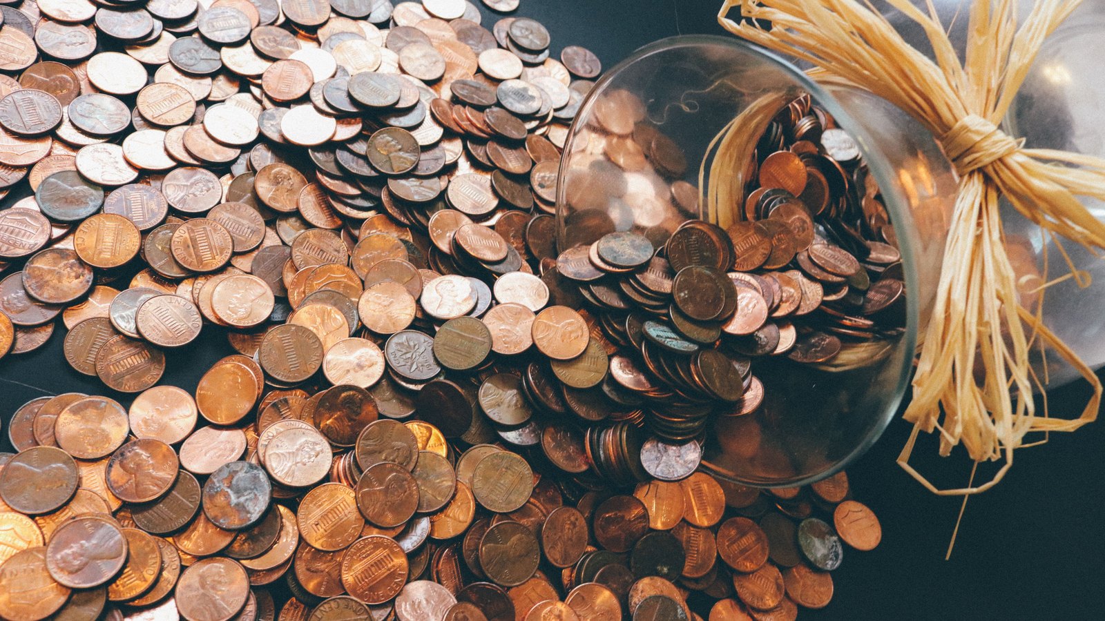 A glass jar tipped on its side on a dark surface with United States one cent coins spilling out and covering the frame, the Lincoln portrait and the words ONE CENT legible on many of them.