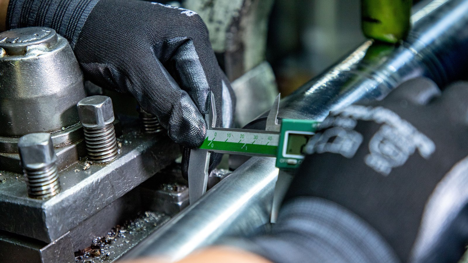 A close photograph of a gloved hand holding a digital vernier caliper across a polished steel shaft clamped in a machine, with metal swarf scattered on the bed below.