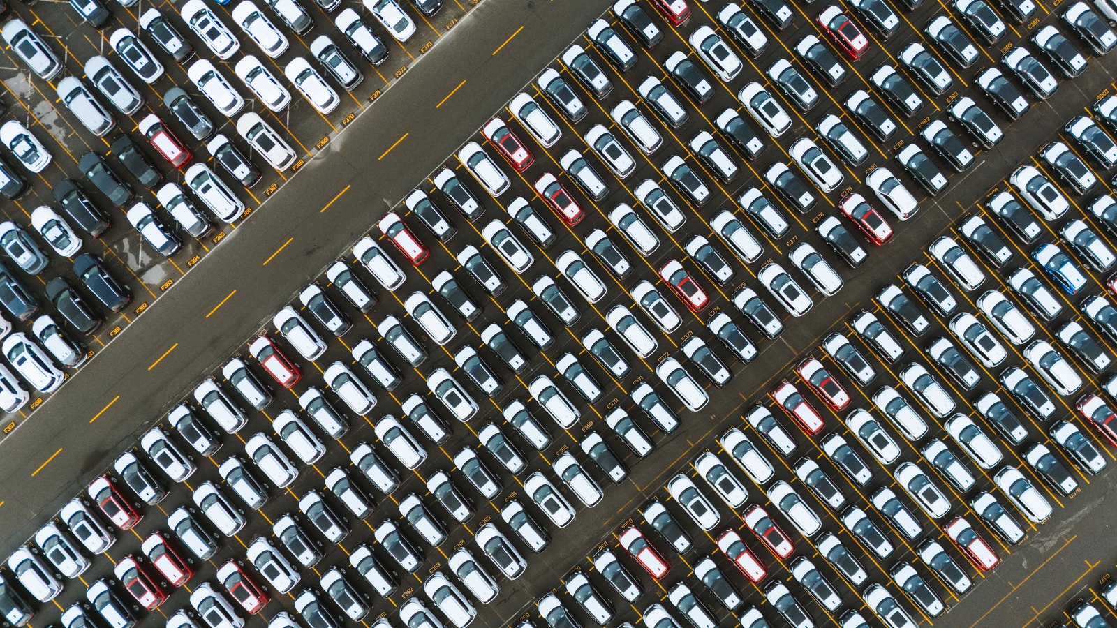 A top-down aerial view of a vehicle compound: several hundred new cars in white, grey, black and red parked nose to tail in long diagonal rows divided by yellow bay lines, with an empty service lane running across the upper left and small painted bay reference codes on the tarmac between the rows.