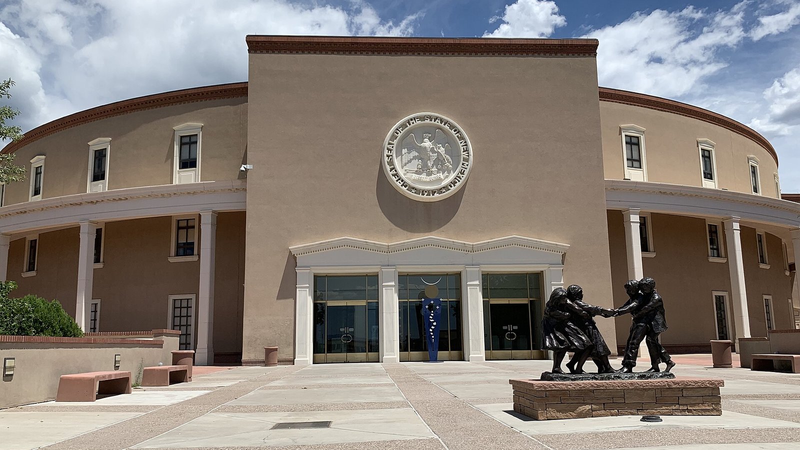 The entrance of the New Mexico State Capitol in Santa Fe, a round tan stucco building with the Great Seal of the State of New Mexico above glass doors and a bronze sculpture of children in the plaza, under a blue sky with clouds.