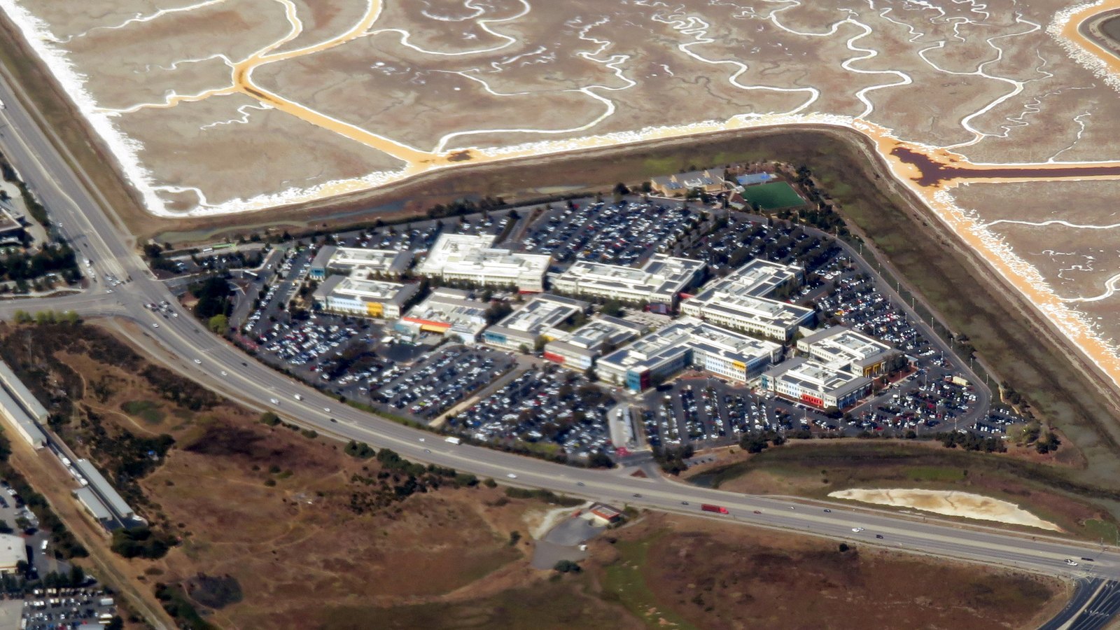An aerial view of a low-rise office campus of white-roofed buildings ringed by full car parks, bounded by a highway and bordered by tidal salt marsh channels on the bay side.