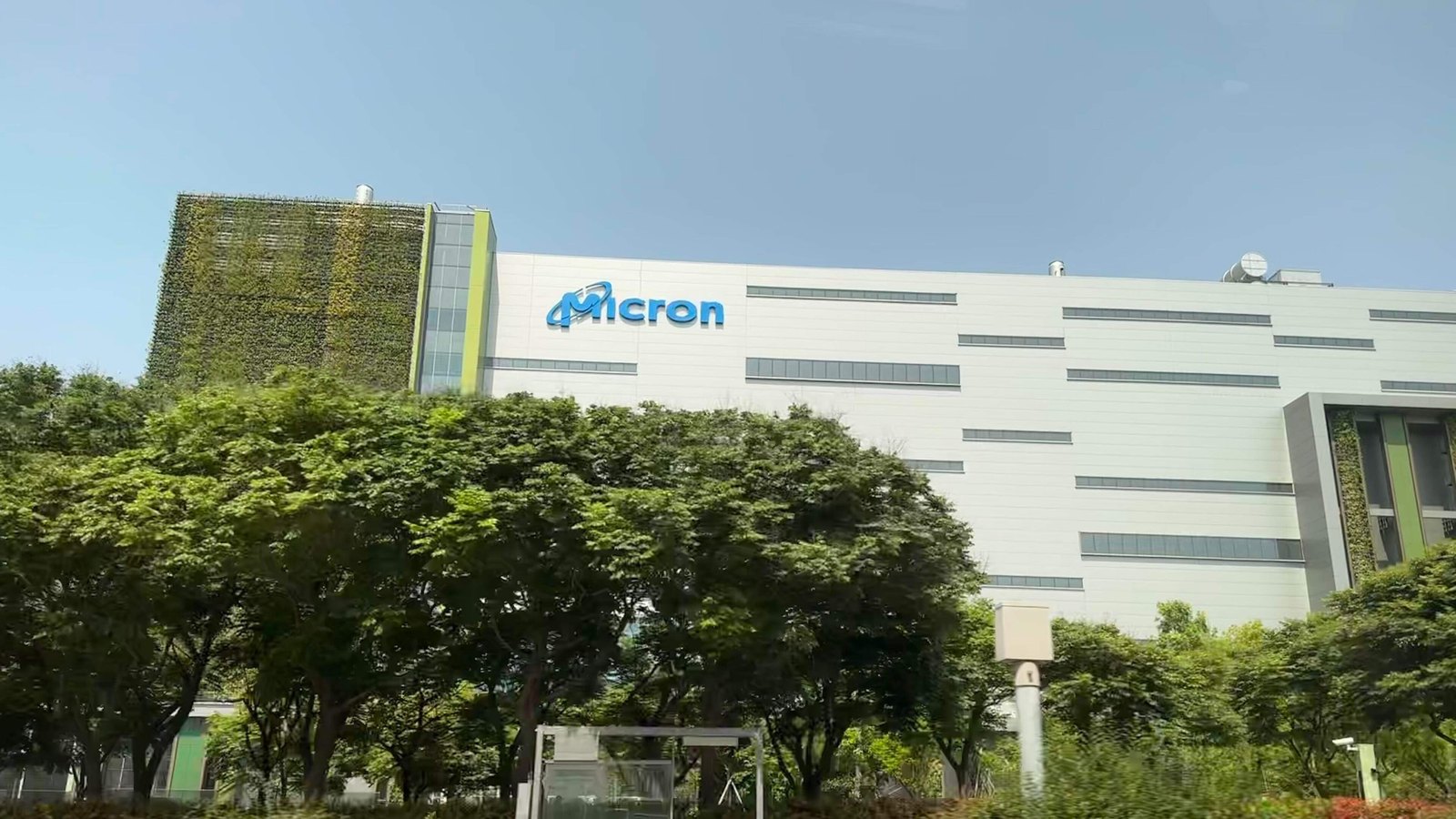 A Micron Technology plant in Taichung, Taiwan: a long white clad building with the company's blue logo mounted high on the facade above rows of narrow windows, seen from the road behind a screen of trees under a clear sky, with a green living wall on the block at the left.
