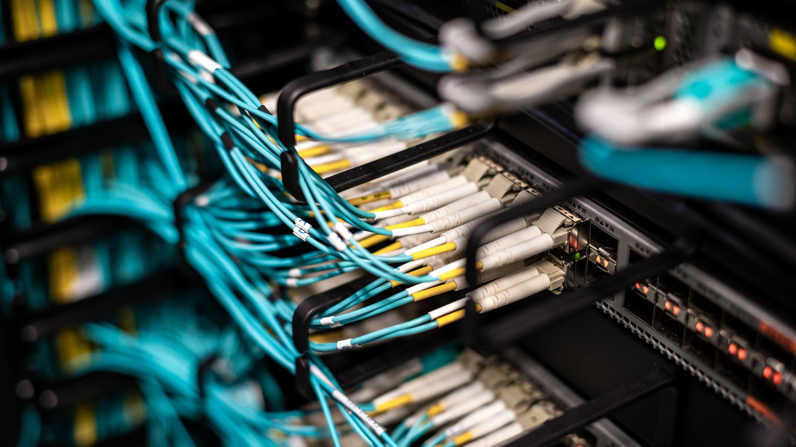 Aqua fibre-optic cables with white connectors plugged into a patch panel in a data centre rack, with small status lights lit.