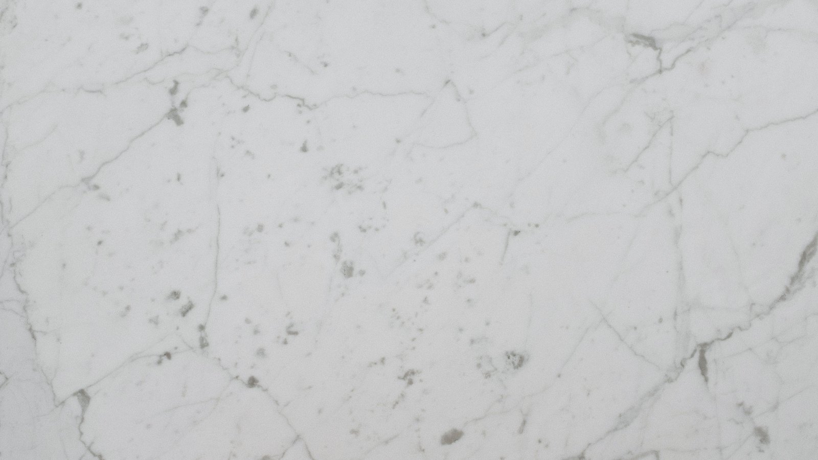 A flat close photograph of a white marble surface crossed by fine grey veins and small darker flecks, filling the frame, with no text, people or setting in view.