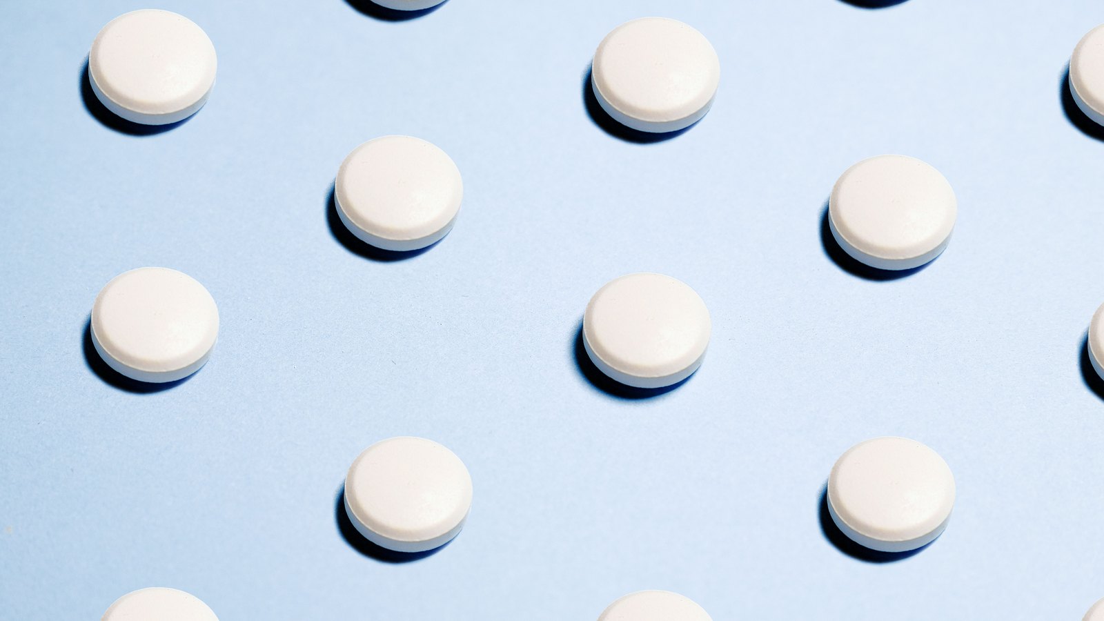 Plain white round tablets laid out in evenly spaced rows on a pale blue surface, each casting a short shadow.