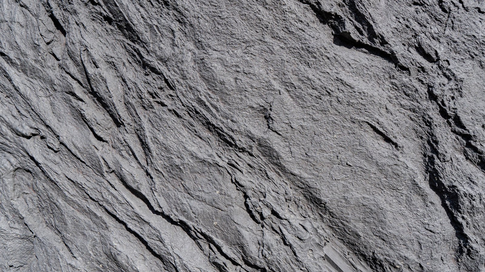A close photograph of a bare grey rock face filling the whole frame, split by diagonal fracture lines running from the lower left toward the upper right, with darker shadowed crevices along the right edge. No text, markings, people or objects are in view.