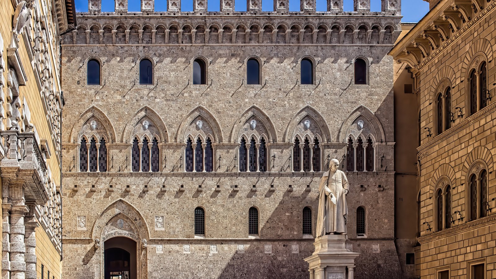 The stone front of Palazzo Salimbeni in Siena, headquarters of Banca Monte dei Paschi di Siena, with a crenellated roofline, rows of pointed arched windows, the name Monte dei Paschi cut above the entrance arch, and a marble statue on a plinth standing in front of it.