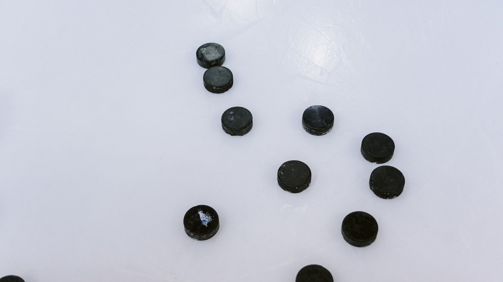 Nine black hockey pucks scattered across a white ice rink.