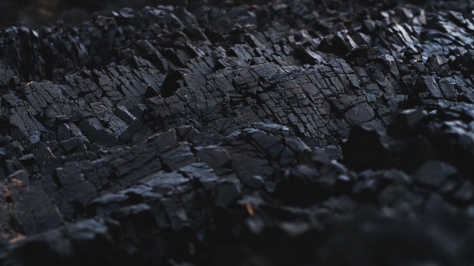 A close photograph of a mass of black mineral filling the frame, broken into sharp blocky facets that catch a faint warm light along their upper edges, the lower right falling away into shadow and nothing else in shot.