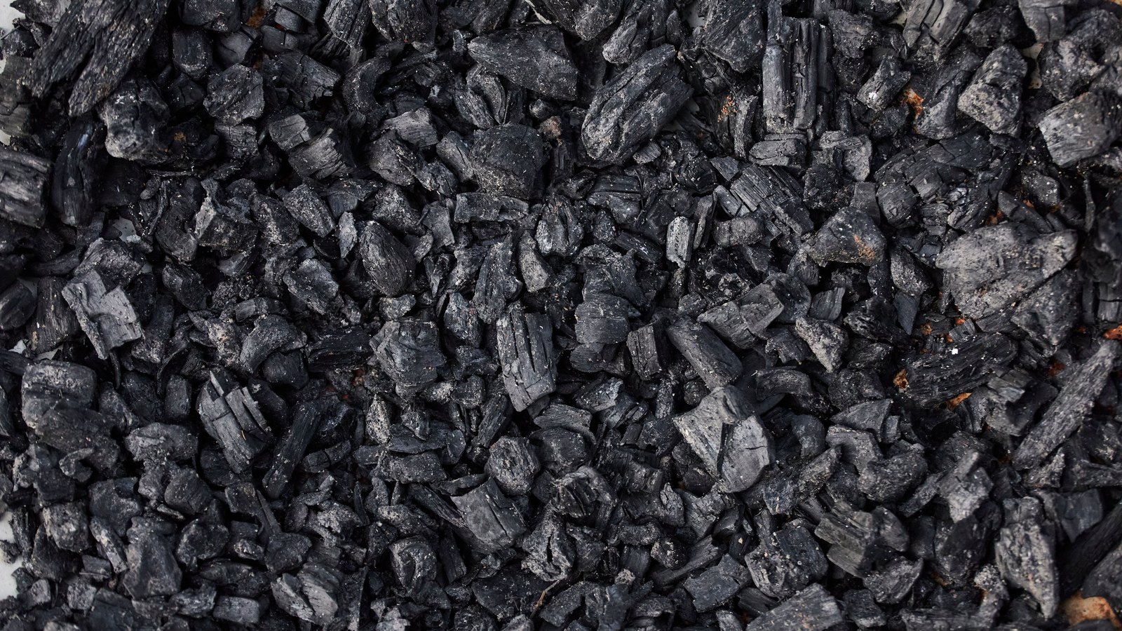 A dense bed of small angular black charcoal fragments photographed from directly above and filling the whole frame, matt black and dark grey with a few dusty pale faces and one or two rust coloured specks. No tool, hand, machine or lettering is in the picture.