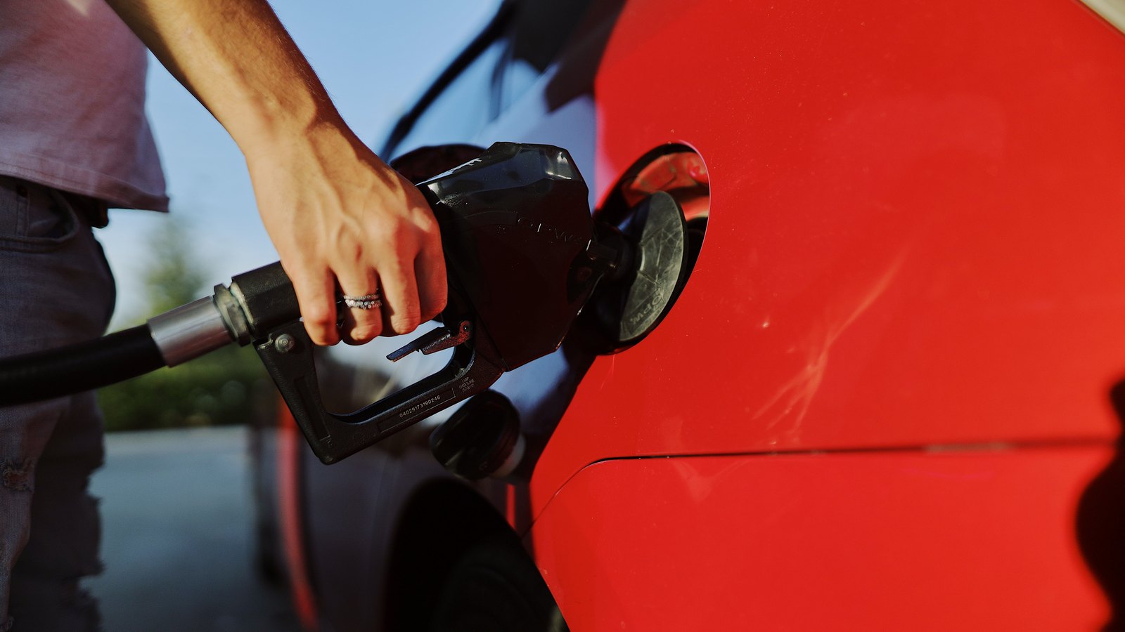 A hand grips a black fuel nozzle inserted into the open filler neck of a red car at a service station, the hose running out of frame to the left, with green foliage and pale sky behind. No face, number plate, signage or brand mark is in view. Illustrative only, not a photograph of this event.