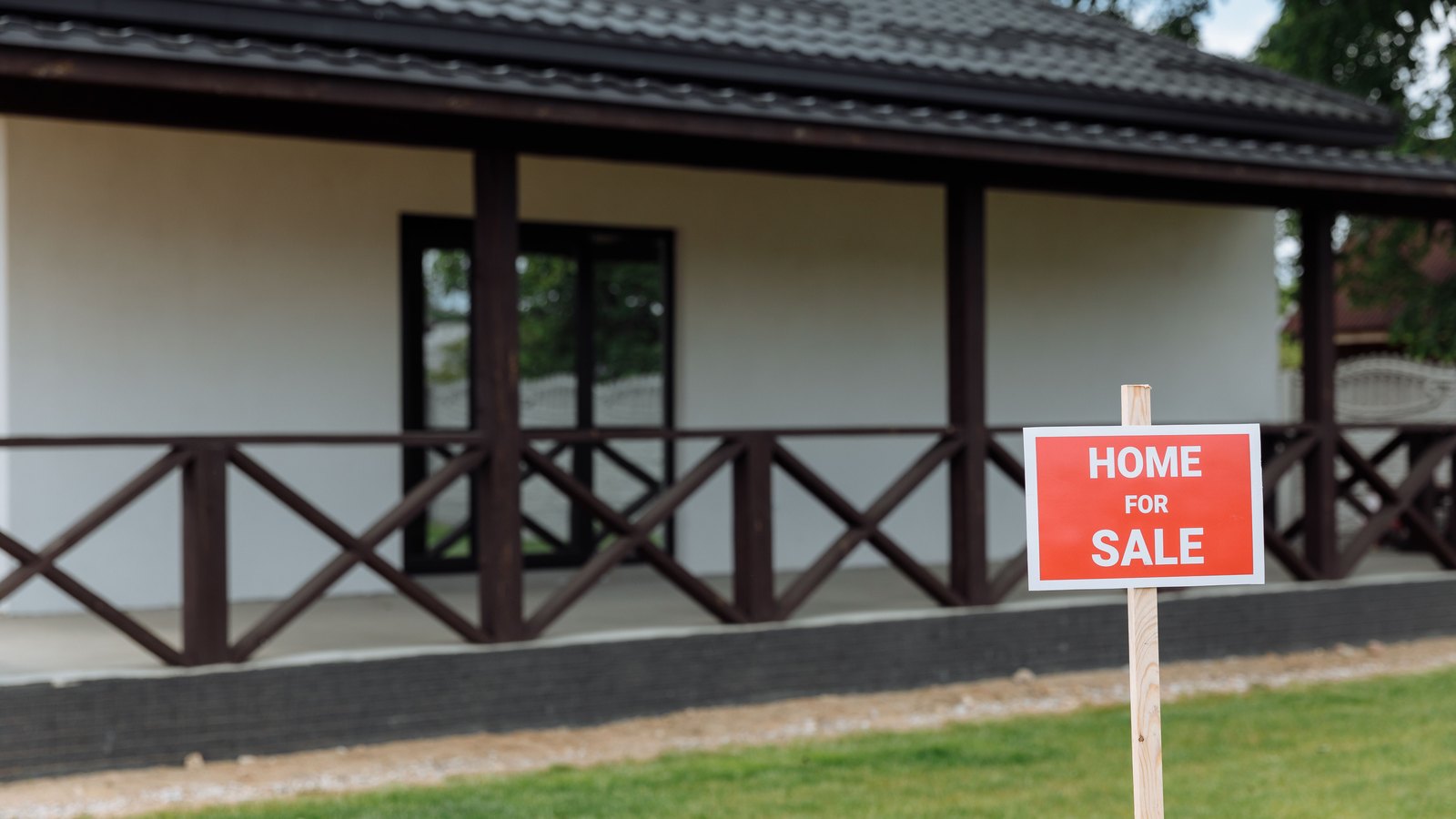 A small red sign reading HOME FOR SALE in white lettering, stapled to a plain wooden stake in a green lawn and sharp in the foreground. Behind it and out of focus stands a single storey house with a dark tiled hip roof, a covered porch and a low wooden rail. No person, no street number, no agency name and no vehicle appears in the frame. Stock photo, not a photograph of this event.
