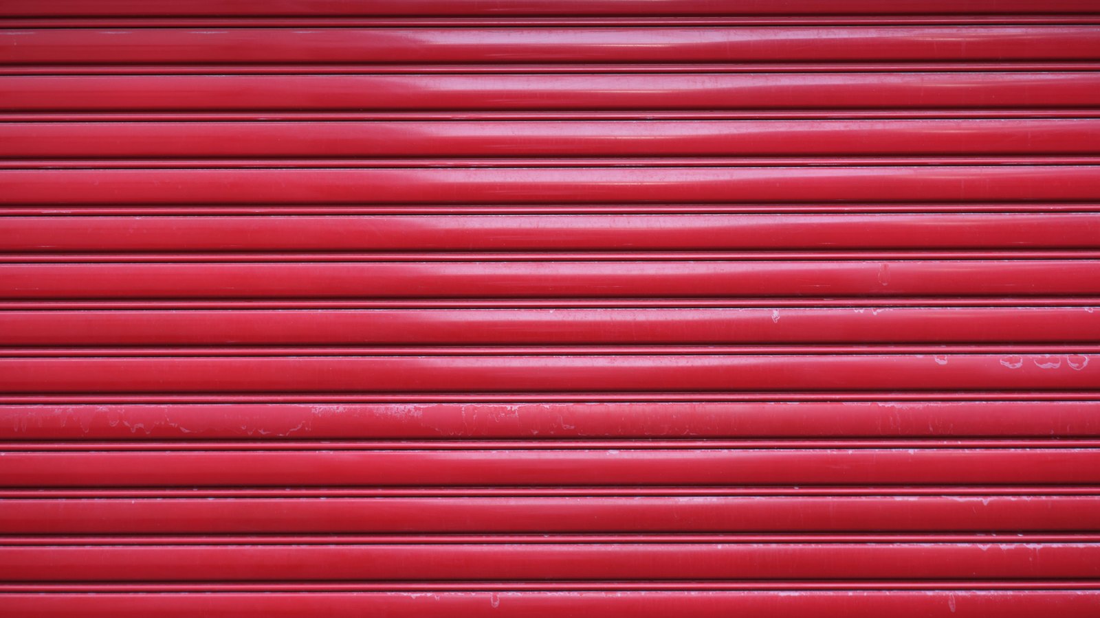 A red painted metal roll-up door filling the frame, its horizontal slats catching flat daylight, the kind fitted to a single industrial unit.