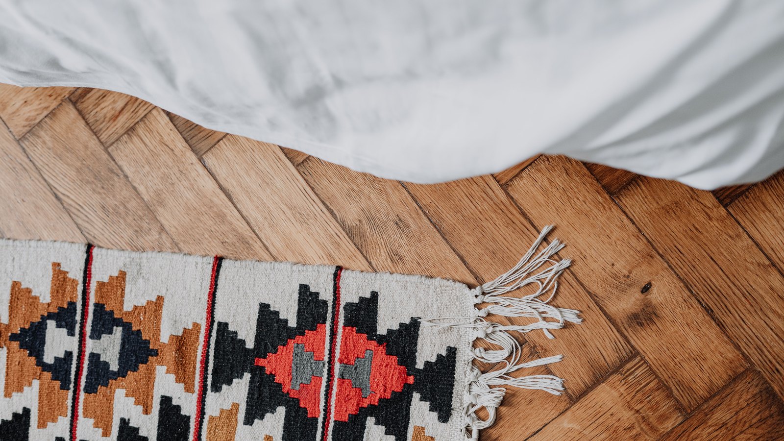 Warm oak herringbone parquet flooring seen from above, with the fringed corner of a patterned kilim rug at the left and the edge of white bedding along the top.
