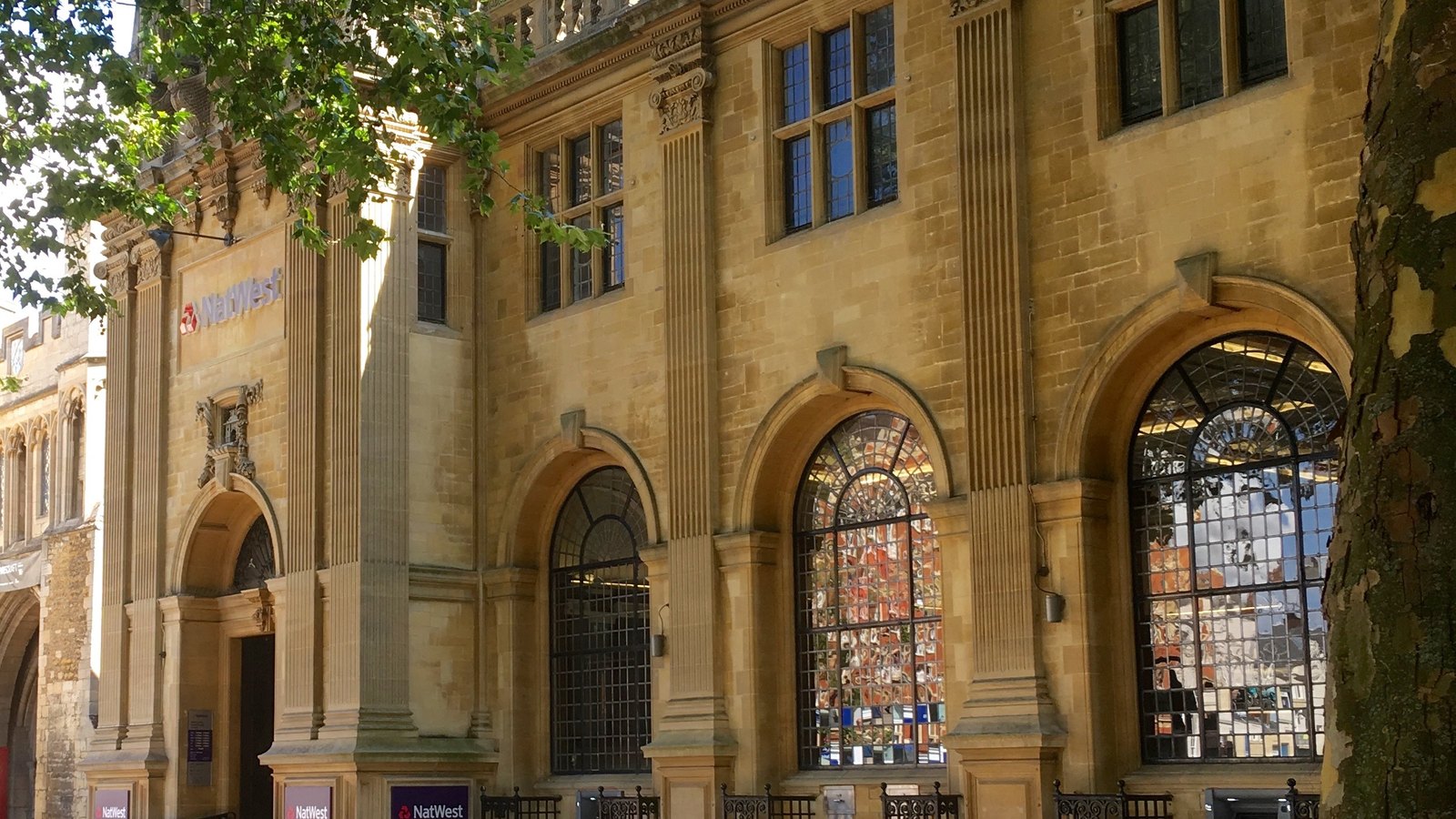 A stone bank building in Peterborough, England, photographed from the pavement at an angle: a honey coloured elevation with fluted pilasters and tall arched leaded windows, the NatWest name and logo mounted on the wall above the entrance and repeated on panels at street level, with a plane tree at the right edge of the frame.