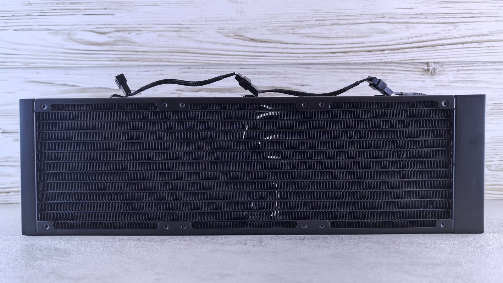 A black liquid cooling radiator photographed straight on against a whitewashed board wall, its dense rows of folded metal fins filling the middle of the frame, with three unmarked fan cables looped across the top edge and no lettering anywhere on it.