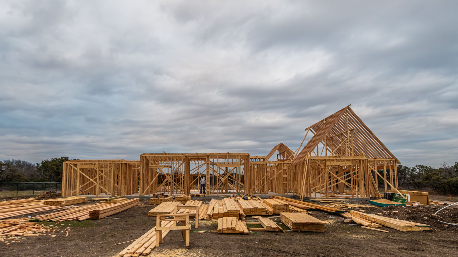 The bare wooden frame of a long single storey house stands on flat cleared ground under a grey overcast sky, with stacks of framing lumber, a sawhorse and loose boards spread across the dirt in front of it, a low scrubby tree line behind, and one small distant unidentifiable figure in a door opening. No signage, lettering or landmarks appear. A generic illustration of new home construction, not a photograph of this event.