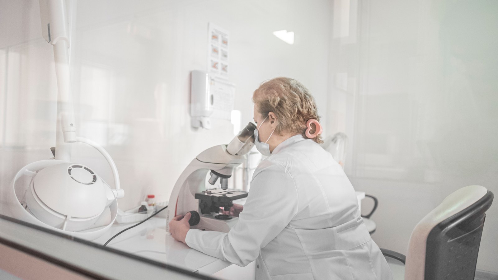 A scientist in a white lab coat examines a sample through a microscope in a bright laboratory.