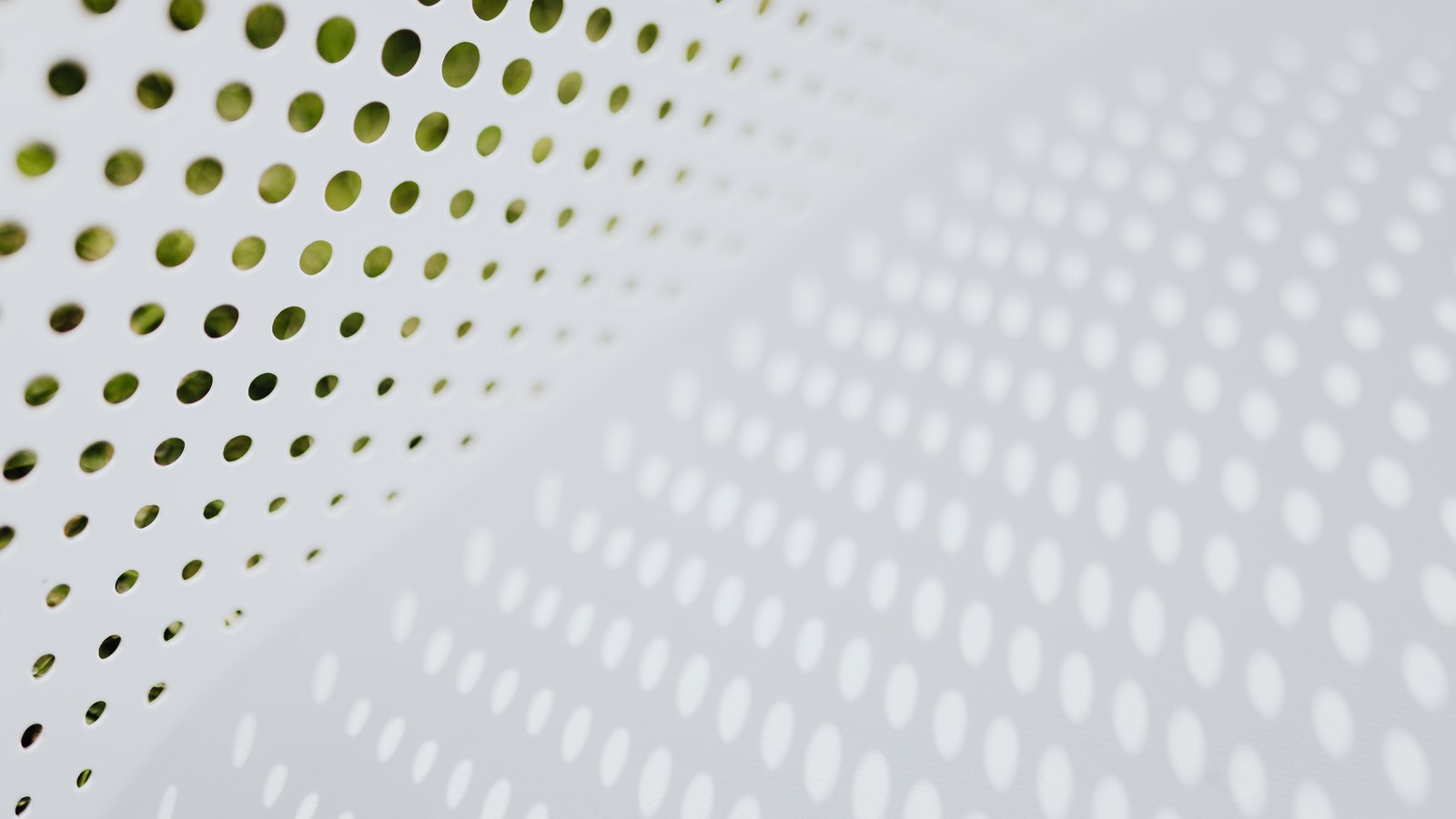 A white panel pierced by a regular grid of round holes, photographed at a shallow angle so the rows recede and blur towards the right, with green foliage showing through the holes along the left edge and the same pattern of dots falling as a soft shadow across the lower half of the frame.