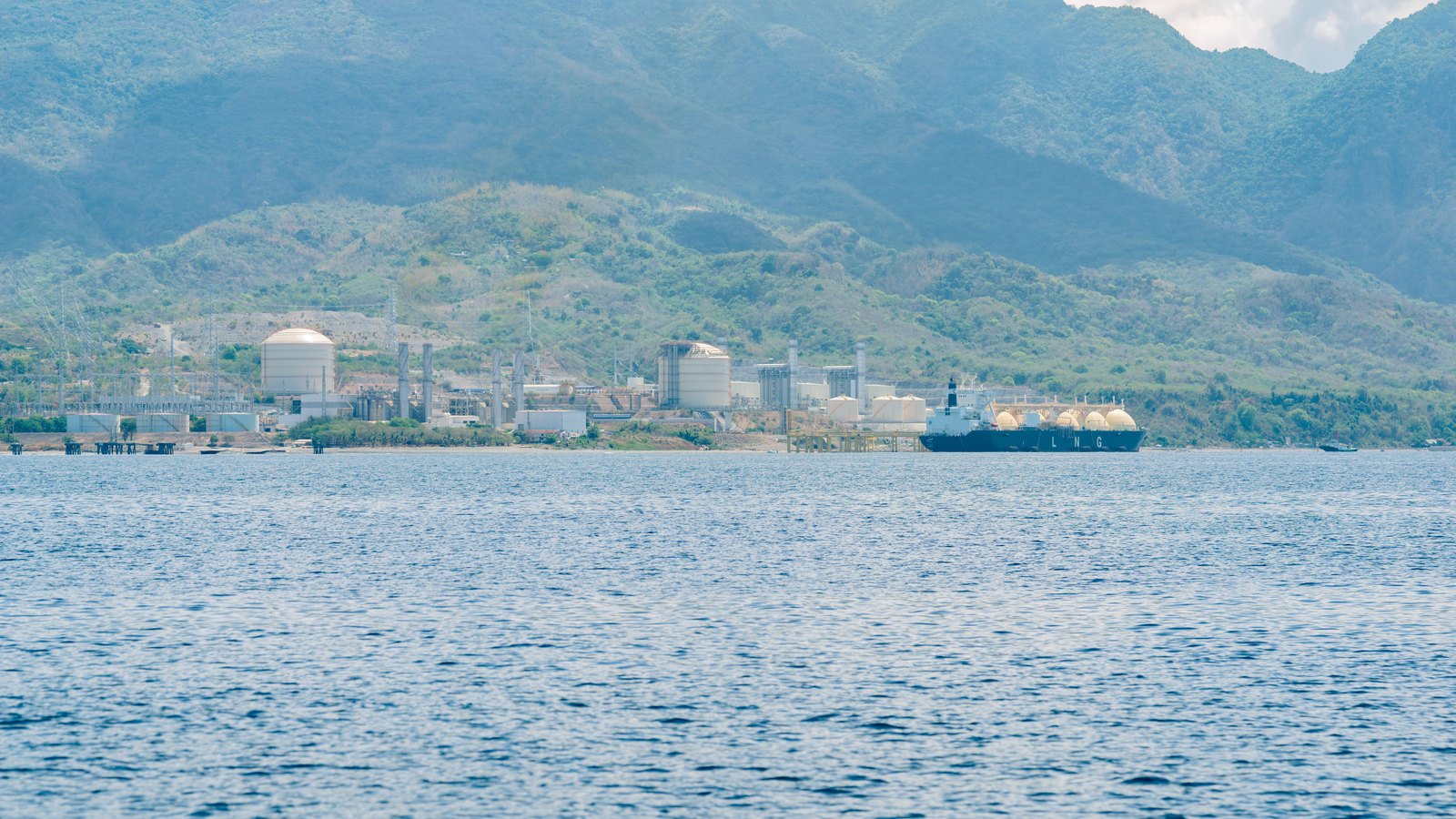 A liquefied natural gas import terminal seen across open water, storage spheres and tanks along the shore below forested hills, and a carrier moored at the jetty with its row of domed cargo tanks above a dark hull carrying the letters L N G in white.