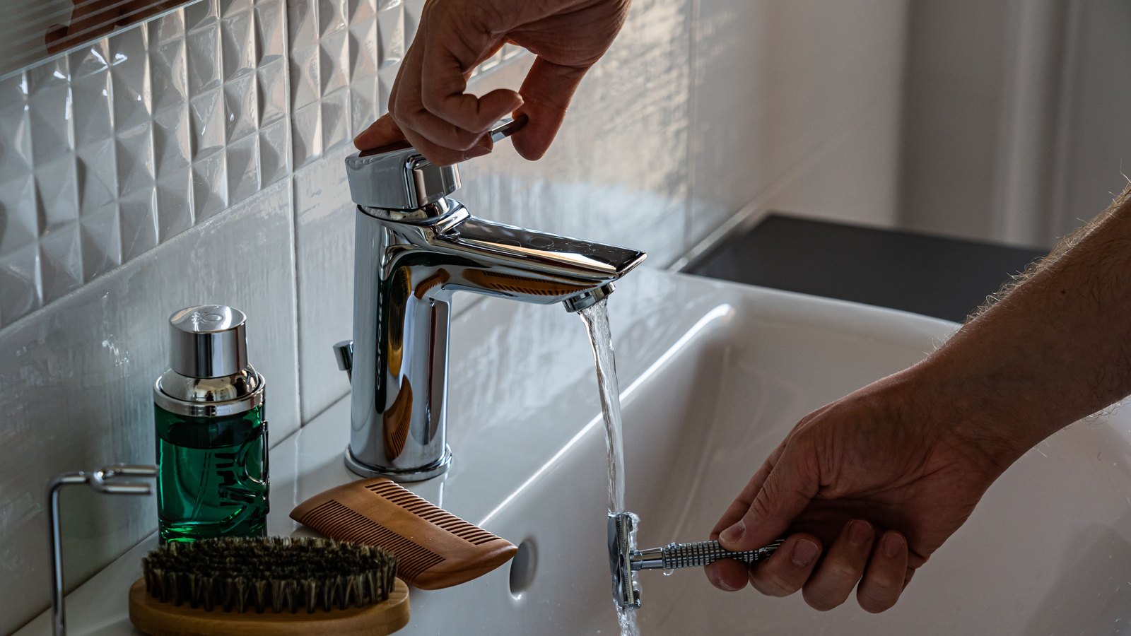 A person turns on a bathroom faucet while holding a razor under the running water.