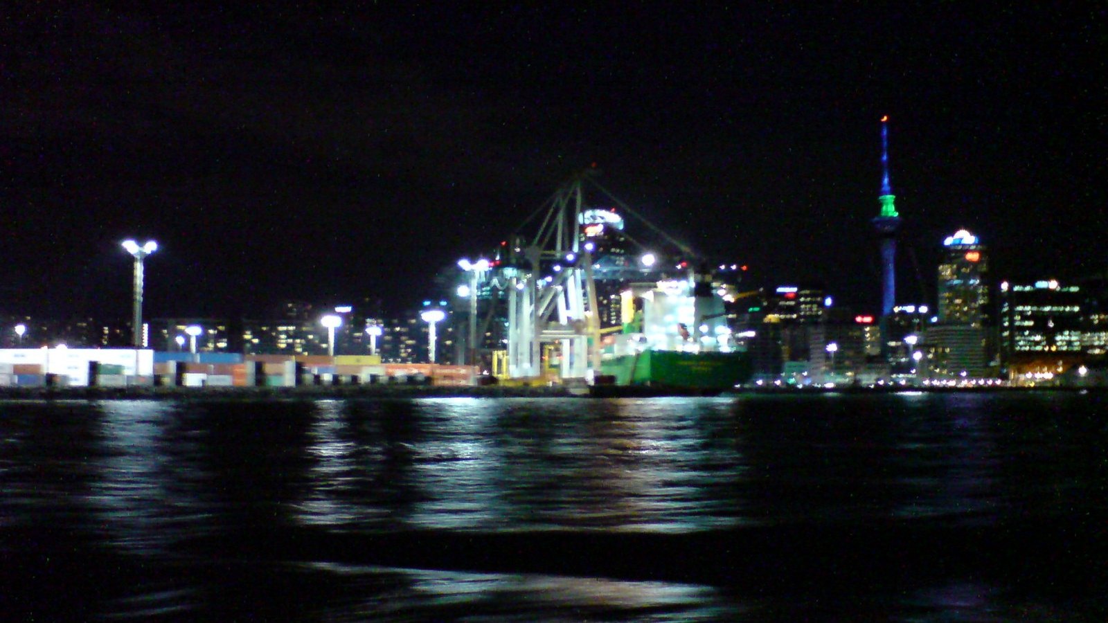 Container cranes operate beside the Auckland skyline at night.